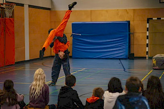 In einer Turnhalle hebt eine Person in dunkler Kleidung eine zweite Person im orangefarbenen Overall kopfüber hoch. Deren Beine sind zu einem Spagat gespreizt. Mehrere Kinder sitzen im Vordergrund auf dem Boden und beobachten die Vorführung aufmerksam.