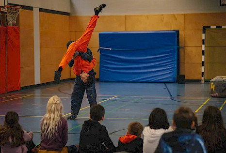 In einer Turnhalle hebt eine Person in dunkler Kleidung eine zweite Person im orangefarbenen Overall kopfüber hoch. Deren Beine sind zu einem Spagat gespreizt. Mehrere Kinder sitzen im Vordergrund auf dem Boden und beobachten die Vorführung aufmerksam.