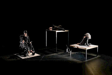 Three performers dressed in black on a dark stage. On the left, a person is seated in a wheelchair on a low platform. In the center, another person lies on a high table. On the right, the third person sits on a low table, with their back turned. Spot lighting highlights their bodies against a completely black background.