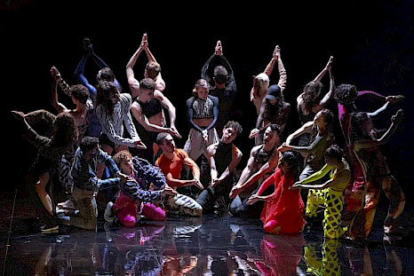 On a dark stage with a reflective floor, twenty-one dancers in brightly colored costumes form a tight semicircle. Standing and kneeling at different levels, they extend their arms toward a shared central point, creating a geometric pattern with their hands under overhead light.