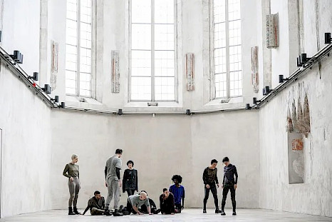 Ten dancers are in a bright white space with high ceilings, like an old church. On the left, a standing performer watches five others sitting and kneeling on the floor. They are looking at four more: two standing to their right, one in front of the group with their back to the camera, and another in the background facing forward.