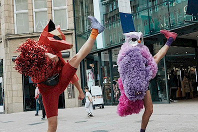 On a city street, two dancers, both with their right leg extended toward the sky and their left leg planted firmly on the ground. The performer on the left is dressed in red with a red fox mask, holding red pompoms with their right arm. The performer on the right wears a pink costume adorned with feathers and a silver owl mask.