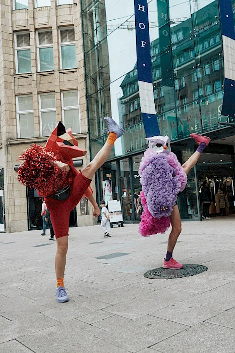 On a city street, two dancers, both with their right leg extended toward the sky and their left leg planted firmly on the ground. The performer on the left is dressed in red with a red fox mask, holding red pompoms with their right arm. The performer on the right wears a pink costume adorned with feathers and a silver owl mask.