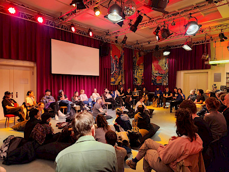 Ungefähr fünfzig Personen sitzen in einem Saal teils auf Stühlen, teils auf Sitzkissen und diskutieren.