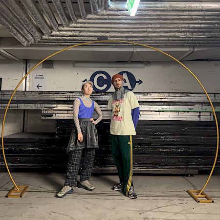 In a dance studio with white walls and a gray floor, two people are dancing. Both stand with their legs apart, one leg bent in front and the other extended back. Their torsos lean backward and their arms are open to the sides.