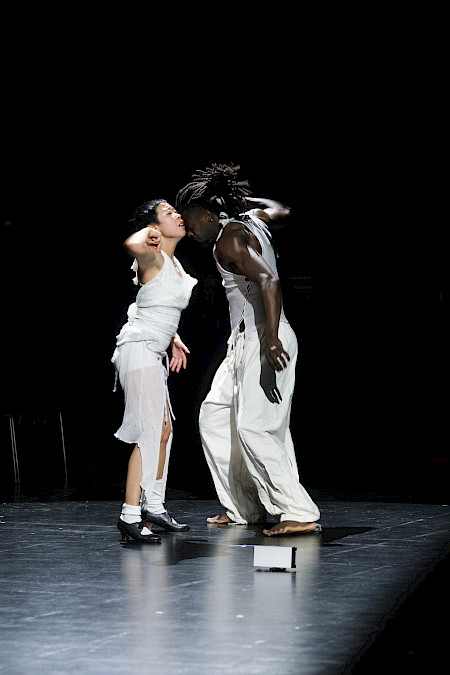 On the left, in black and white, a person on a stage opens and extends their arms, with one leg crossed in front of the other. They wear a light sleeveless top and dark trousers. On the right, in color, a person tilts their torso to one side with both arms extended upward. They wear a light sleeveless top and loose trousers.