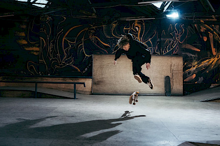 A skateboarder dressed in black floats in the air performing a trick inside an indoor skatepark. The skateboard spins beneath his feet as a strong light casts his elongated shadow on the concrete floor. In the background, dark graffiti-covered walls, ramps, and rails complete the scene.