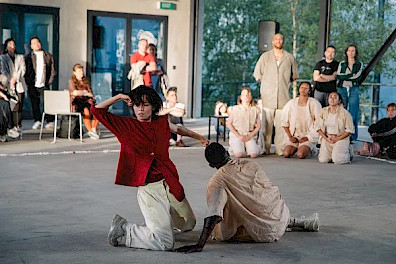 Two dancers perform on a concrete floor, while four more observe from the back. They are surrounded by an audience. In the foreground, a person in a red shirt and light-colored pants kneels with open hands and an intense expression. Beside them, another dancer dressed in beige sits on the floor, leaning on one hand with their body turned away. Large windows reveal green trees outside, and a speaker on a stand is visible near the wall.