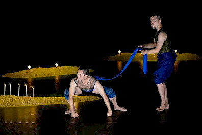 Against a black background, three small golden sand mounds emerge along a diagonal line stretching from the front right of the stage to the back left. In front of them two performers dressed in blue stand: one upright, holding a blue fabric tied to the back of his partner, who crouches about half a meter away, legs bent and fists pressed firmly against the floor.