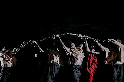 Six dancers stand side by side in a straight line, holding a wooden pole together above their heads. Two are dressed completely in red, while the other four wear beige shirts paired with wide gray trousers. Light falls from above, casting strong shadows across their bodies, while the background remains black.