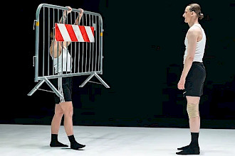 Auf einer Bühne mit weißem Boden und schwarzen Wänden stehen sich zwei Tänzer:innen gegenüber. Beide tragen schwarze Strümpfe, schwarze Shorts und weiße, ärmellose Hemden. Die performende Person auf der linken Seite hält eine Metallbarriere mit einem rot-weißen Streifen, der daran hängt. Ihr gegenüber, auf der rechten Seite des Bildes, steht die zweite performende Person und sieht sie aufmerksam an.