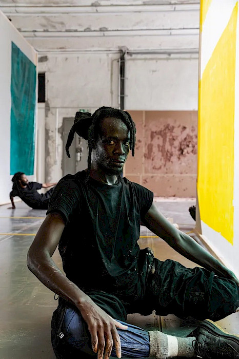 A person with tied-up hair sits sideways on the floor of a bright industrial-looking room, with one leg bent and the other extended forward, looking directly at the camera. Large turquoise and yellow fabric panels hang beside them, while another person lies on the floor in the background.