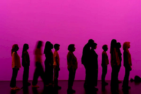 Auf einem pinkfarbenen Hintergrund stehen die Silhouetten von dreizehn Menschen unterschiedlichen Alters (von Teenagern bis zu älteren Erwachsenen) im Profil, alle mit Blickrichtung zur rechten Seite der Bühne.