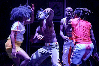 Vier Personen tanzen unter sanftem violettem Licht auf einer dunklen Bühne. Links trägt eine Person eine weiße Bluse und einen gelben Rock, in der Mitte steht eine weitere Person ohne Hemd mit heller Hose und rechts zwei Personen in hellen Hosen, von denen eine ein weißes Tanktop und die andere ein rotes Hemd mit langen Zöpfen trägt, in dynamischen Posen.