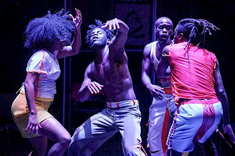 Vier Personen tanzen unter sanftem violettem Licht auf einer dunklen Bühne. Links trägt eine Person eine weiße Bluse und einen gelben Rock, in der Mitte steht eine weitere Person ohne Hemd mit heller Hose und rechts zwei Personen in hellen Hosen, von denen eine ein weißes Tanktop und die andere ein rotes Hemd mit langen Zöpfen trägt, in dynamischen Posen.