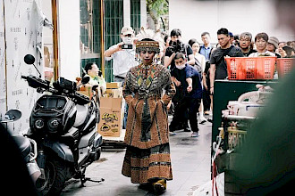 Auf einem belebten Markt oder einer belebten Straße geht eine Person in reich verzierter Kleidung voran. Sie trägt ein langes Kleid mit aufwendigen geometrischen Mustern in Erdtönen, mehrreihige Halsketten und einen kunstvollen Kopfschmuck, der mit Perlen und Federn verziert ist. Ihre Hände sind vor ihr gefaltet, während sie nach unten schaut. Hinter ihr folgt ihr eine Gruppe von Menschen dicht auf den Fersen; einige halten Kameras und Mobiltelefone in den Händen.