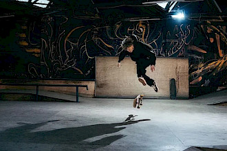 Ein schwarz gekleideter Skateboarder schwebt in der Luft und führt in einem Indoor-Skatepark einen Trick aus. Das Skateboard dreht sich unter seinen Füßen, während ein starkes Licht seinen langgestreckten Schatten auf den Betonboden wirft. Im Hintergrund vervollständigen dunkle, mit Graffiti bedeckte Wände, Rampen und Geländer die Szene.