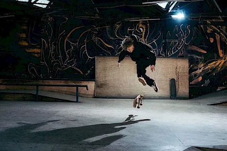 A skateboarder dressed in black floats in the air performing a trick inside an indoor skatepark. The skateboard spins beneath his feet as a strong light casts his elongated shadow on the concrete floor. In the background, dark graffiti-covered walls, ramps, and rails complete the scene.