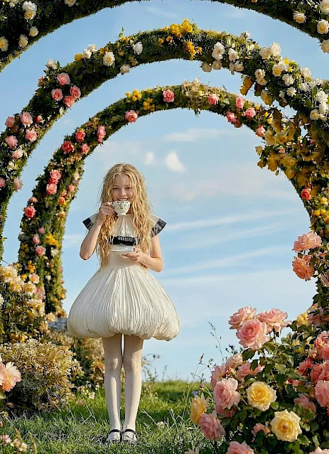 Ein Kind mit langen hellen Haaren steht auf einer Wiese unter mehreren blumenumrankten Bögen und hält eine Tasse mit Untertasse vor sich. Es trägt ein helles, ballonförmiges Kleid, umgeben von rosa, gelben und weißen Blüten.
