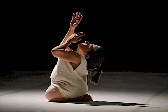 On a stage with a white floor and dark background, a performer sits low to the ground with legs folded beneath the body, wearing a light sleeveless garment. The torso leans slightly back while both arms are raised upward, hands open. Overhead light illuminates the figure and casts a soft shadow on the floor.